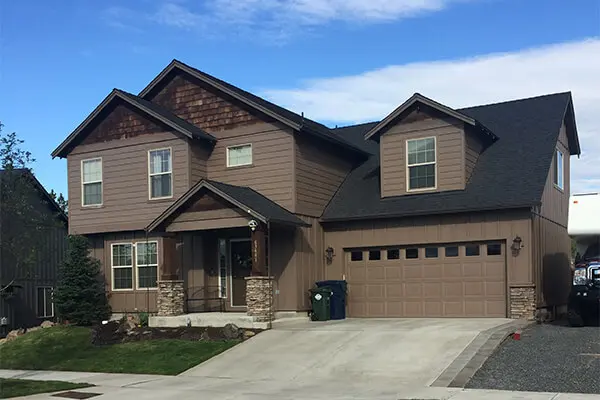 A two-story suburban home with brown siding and stone veneer accents, featuring a paved driveway, a well-maintained lawn, and a garage entrance, captured on a sunny day.