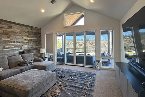 A bright, contemporary living area featuring a floor-to-ceiling stone feature wall, a plush gray sectional sofa, and large glass patio doors that offer a view of the outdoor landscape.