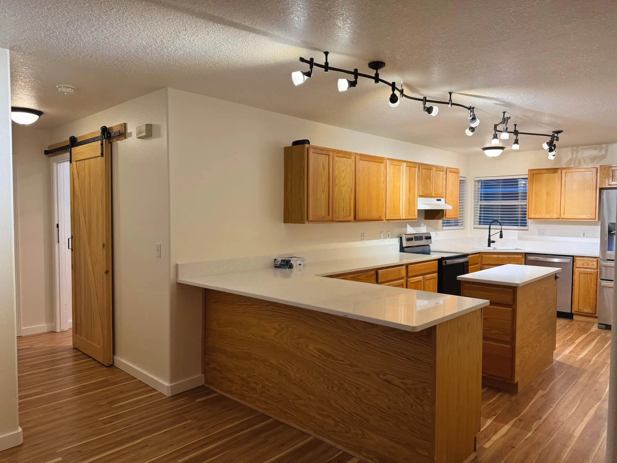 A well-lit, open-concept kitchen featuring light-toned wooden cabinets, a large quartz-topped island with a sink, stainless steel appliances, and sliding barn doors, creating a warm and modern home aesthetic.
