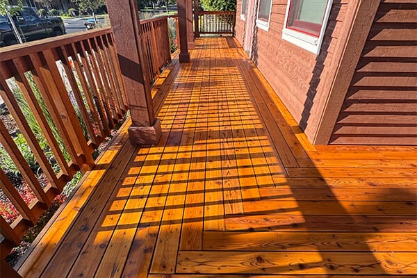 A newly stained, natural wood deck featuring horizontal railing planks and a cozy seating area, overlooking a lush green yard on a sunny day.