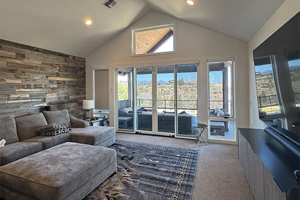 A bright, contemporary living area featuring a floor-to-ceiling stone feature wall, a plush gray sectional sofa, and large glass patio doors that offer a view of the outdoor landscape.