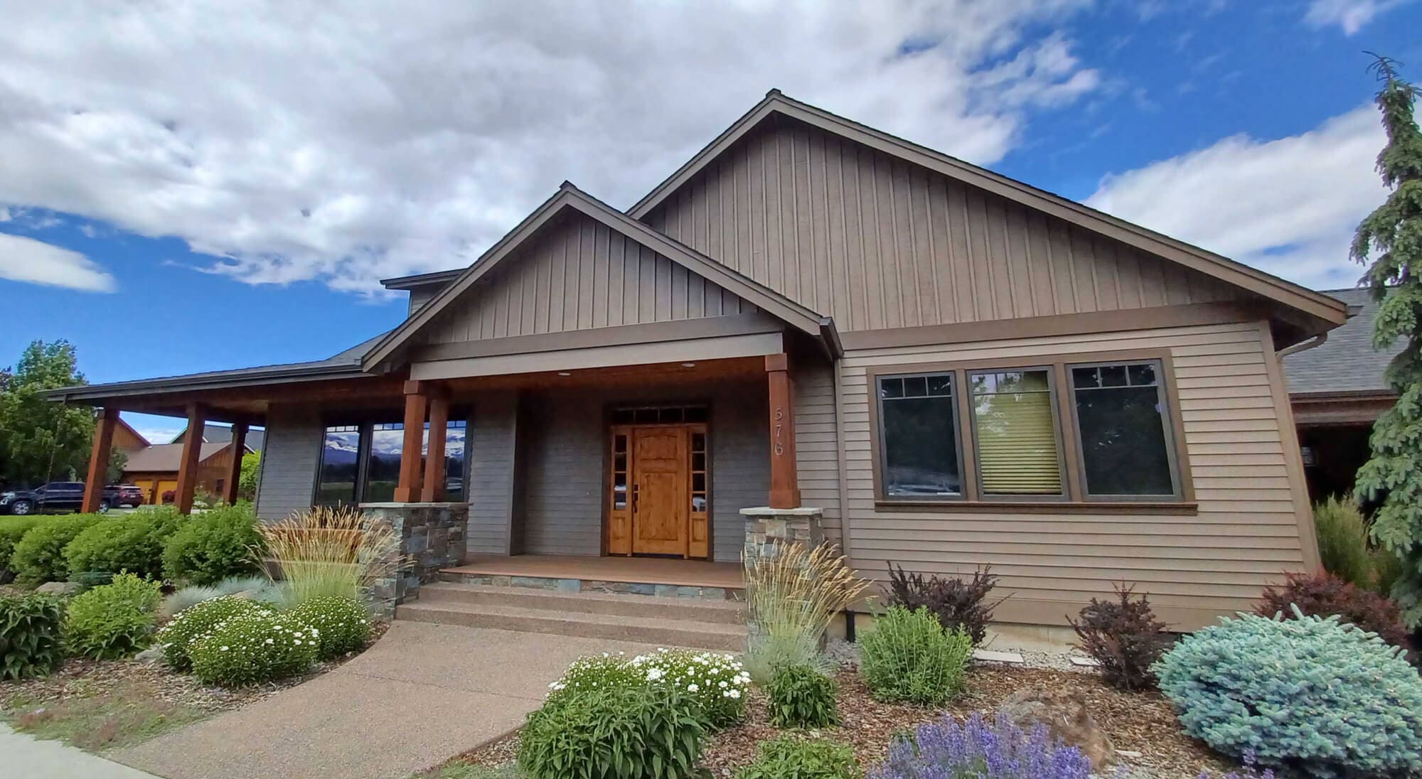 A well-maintained, single-story suburban residence with warm-toned siding, a welcoming front porch supported by stone columns, and manicured landscaping under a bright blue sky.