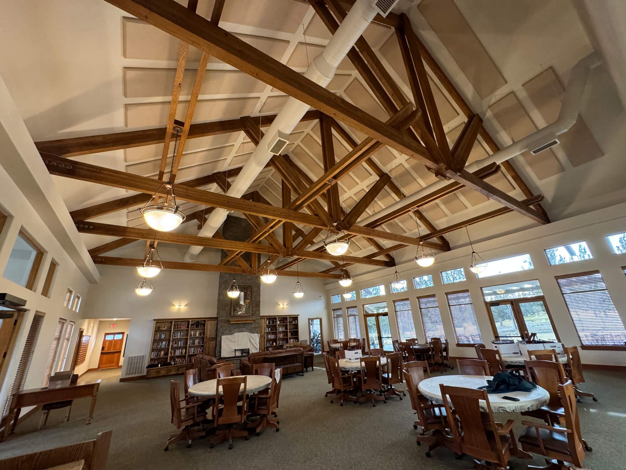 A spacious, rustic-modern great room featuring impressive vaulted ceilings with exposed heavy timber trusses, hardwood floors, a stone fireplace, and a large dining area filled with natural light.