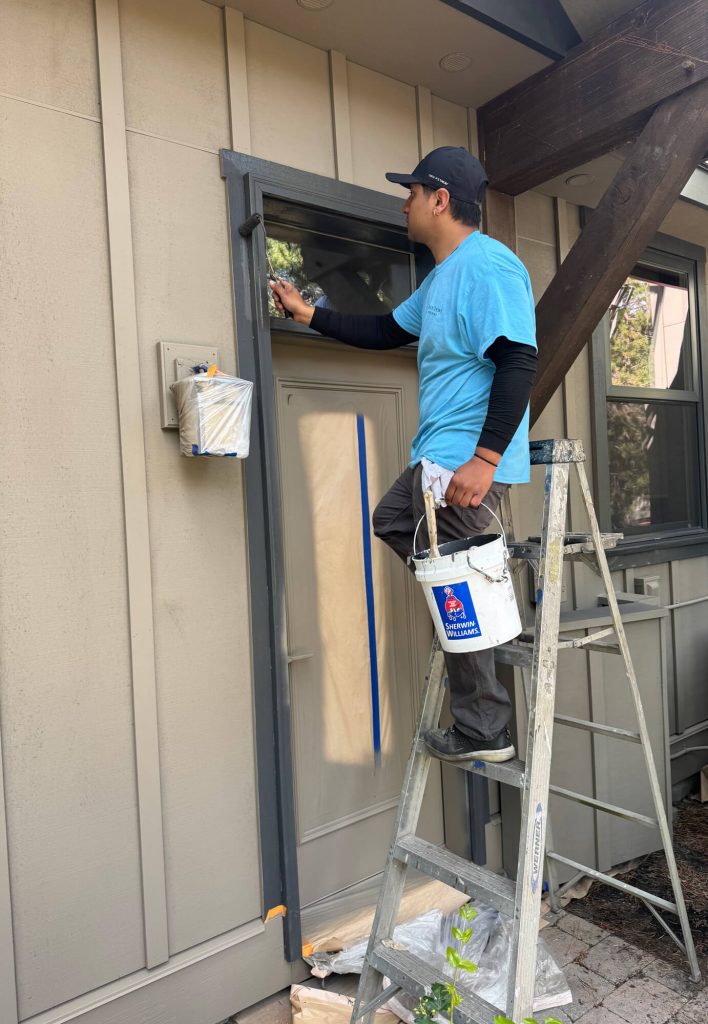 A West Rivers Painting crew member wearing a blue uniform is meticulously applying fresh paint to the exterior door frame of a residential home while using a ladder for access.